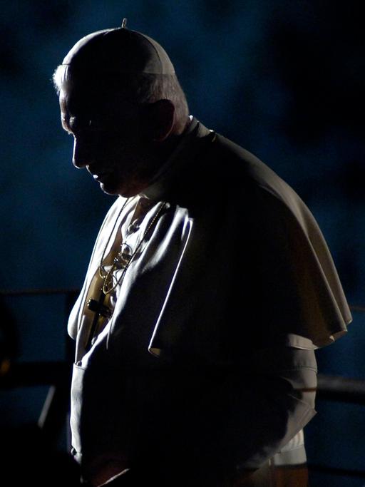 Papst Benedikt XVI. steht in weißer Robe und Kopfbedeckung mit gesenktem Kopf seitlich zur Kamera. Das Licht leuchtet ihn von der Seite an, so dass der größte Teil seines Gesichts verschattet bleibt.