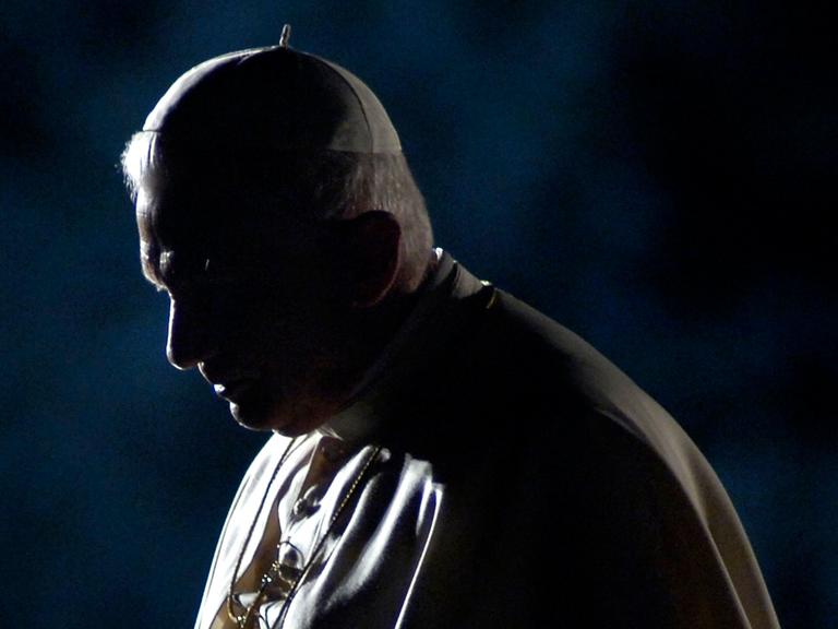 Papst Benedikt XVI. steht in weißer Robe und Kopfbedeckung mit gesenktem Kopf seitlich zur Kamera. Das Licht leuchtet ihn von der Seite an, so dass der größte Teil seines Gesichts verschattet bleibt.