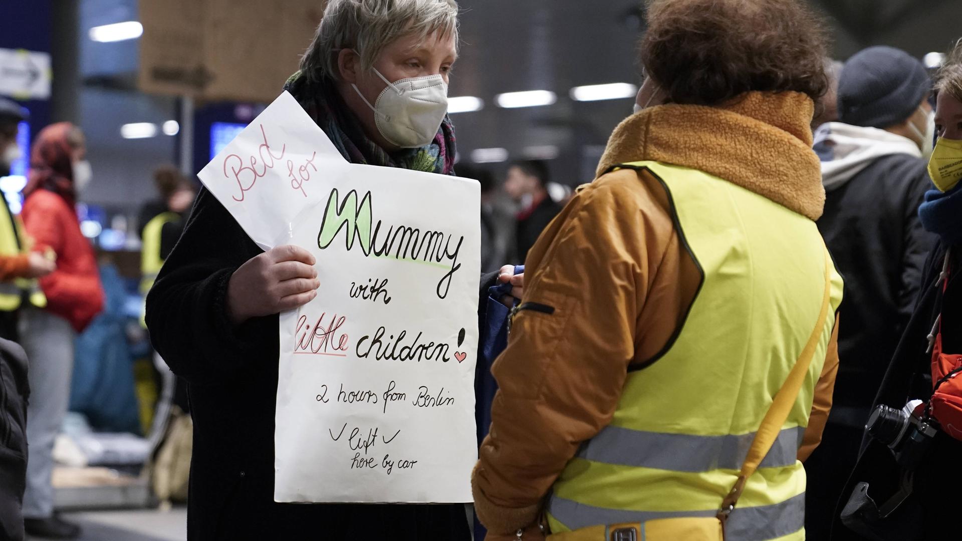 Eine Frau mit Atemschutzmaske trägt ein Schild vor dem Bauch, auf dem sie in englischer Sprache eine Unterkunft für eine Mutter mit kleinen Kindern anbietet. Mit dem Rücken zur Kamera steht vor ihr eine dunkelhäutige Frau, die eine gelbe Warnweste trägt. Eine Frau mit Atemschutzmaske trägt ein Schild vor dem Bauch, auf dem sie in englischer Sprache eine Unterkunft für eine Mutter mit kleinen Kindern anbietet. Mit dem Rücken zur Kamera steht vor ihr eine dunkelhäutige Frau, die eine gelbe Warnweste trägt.