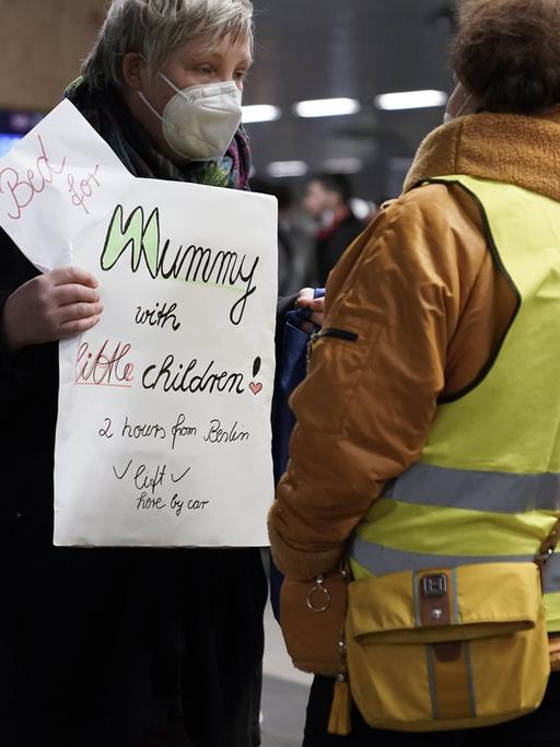 Eine Frau mit Atemschutzmaske trägt ein Schild vor dem Bauch, auf dem sie in englischer Sprache eine Unterkunft für eine Mutter mit kleinen Kindern anbietet. Mit dem Rücken zur Kamera steht vor ihr eine dunkelhäutige Frau, die eine gelbe Warnweste trägt.