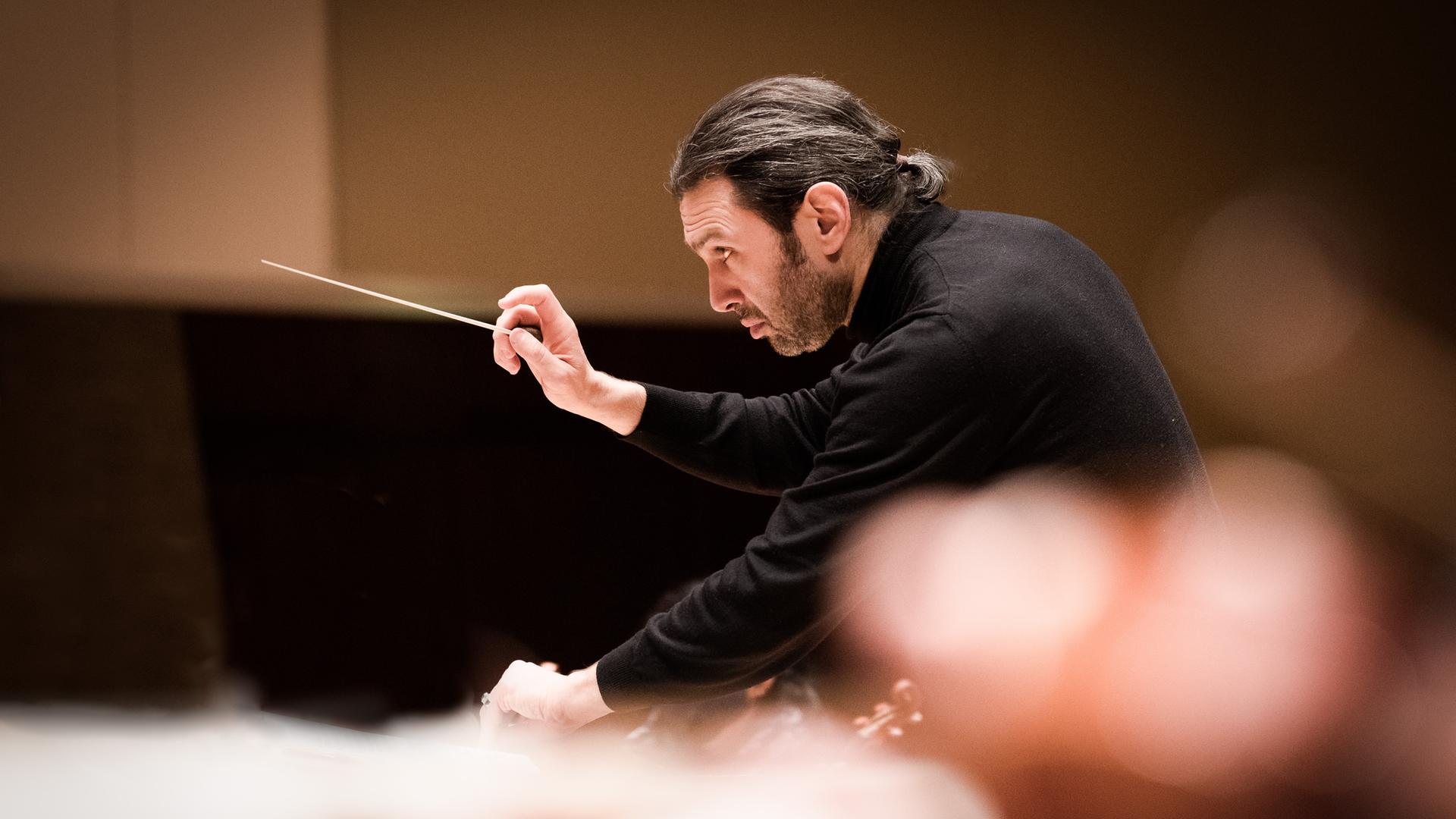 Vladimir Jurowski gibt gebeugt seinen Musikern mit dem Dirigierstab ein Zeichen für den Einsatz.