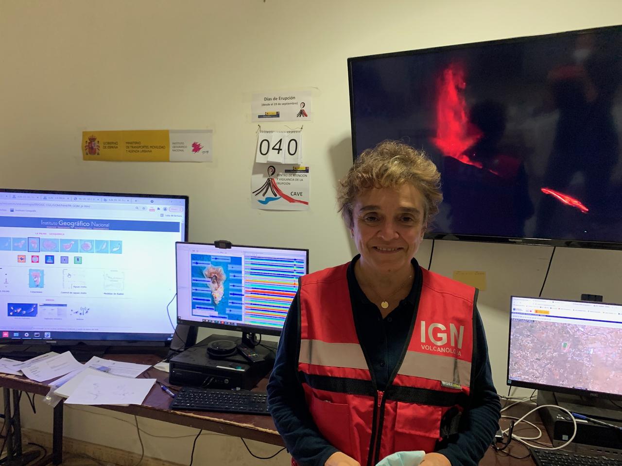Eine ältere Dame mit orangener Signalweste steht in einem Büroraum mit vielen bunten Bildschirmen, von denen einer in Großformat den brennenden Vulkan zeigt. Eine ältere Dame mit orangener Signalweste steht in einem Büroraum mit vielen bunten Bildschirmen, von denen einer in Großformat den brennenden Vulkan zeigt.