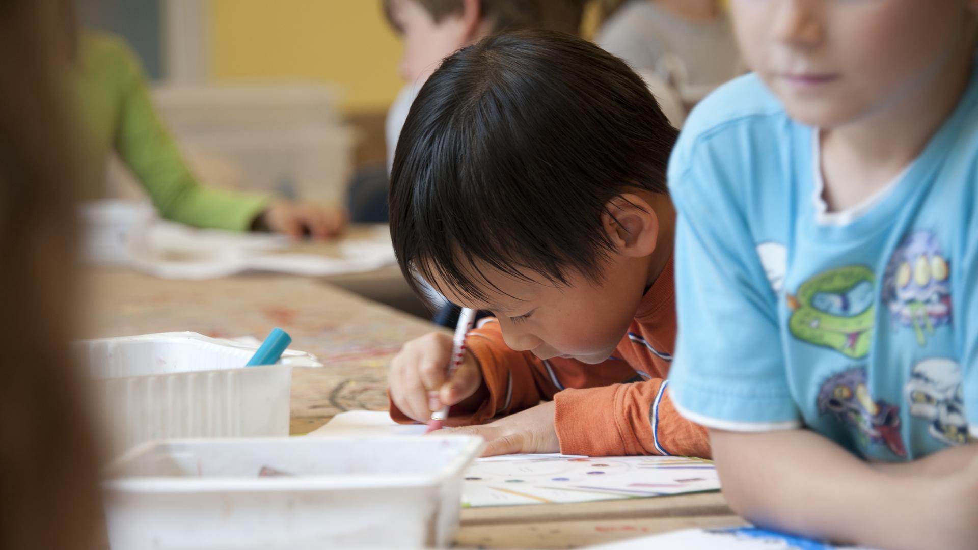 Kinder in einem Klassenzimmer. Im Bildzentrum ist ein Kind mit dunklen Haaren und orangefarbenem Shirt. Es malt. Kinder in einem Klassenzimmer. Im Bildzentrum ist ein Kind mit dunklen Haaren und orangefarbenem Shirt. Es malt.