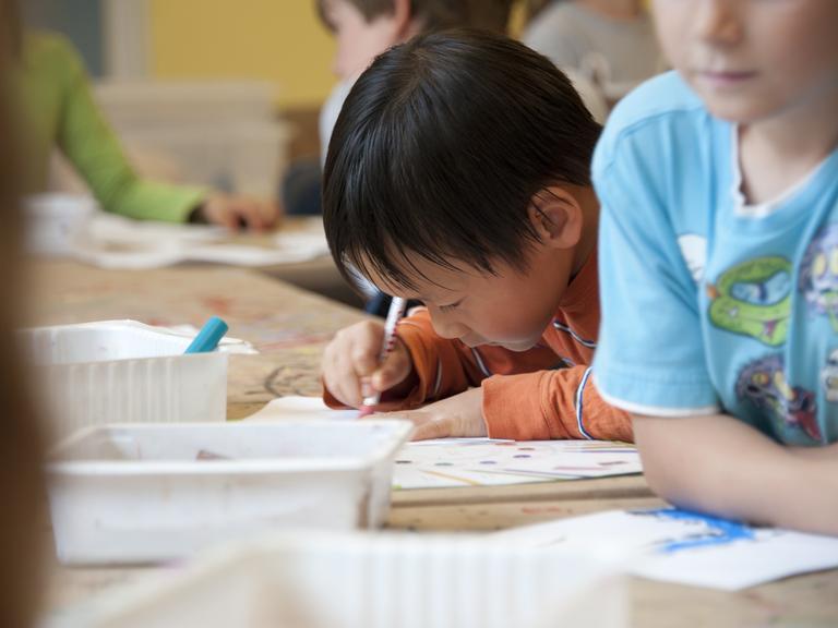 Kinder in einem Klassenzimmer. Im Bildzentrum ist ein Kind mit dunklen Haaren und orangefarbenem Shirt. Es malt.