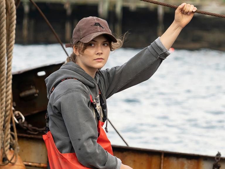Schauspielerin Emilia Jones sitzt in einer Szene des oscarprämierten Films "Coda" in Arbeitskleidung einem Fischerboot und schaut in die Kamera.