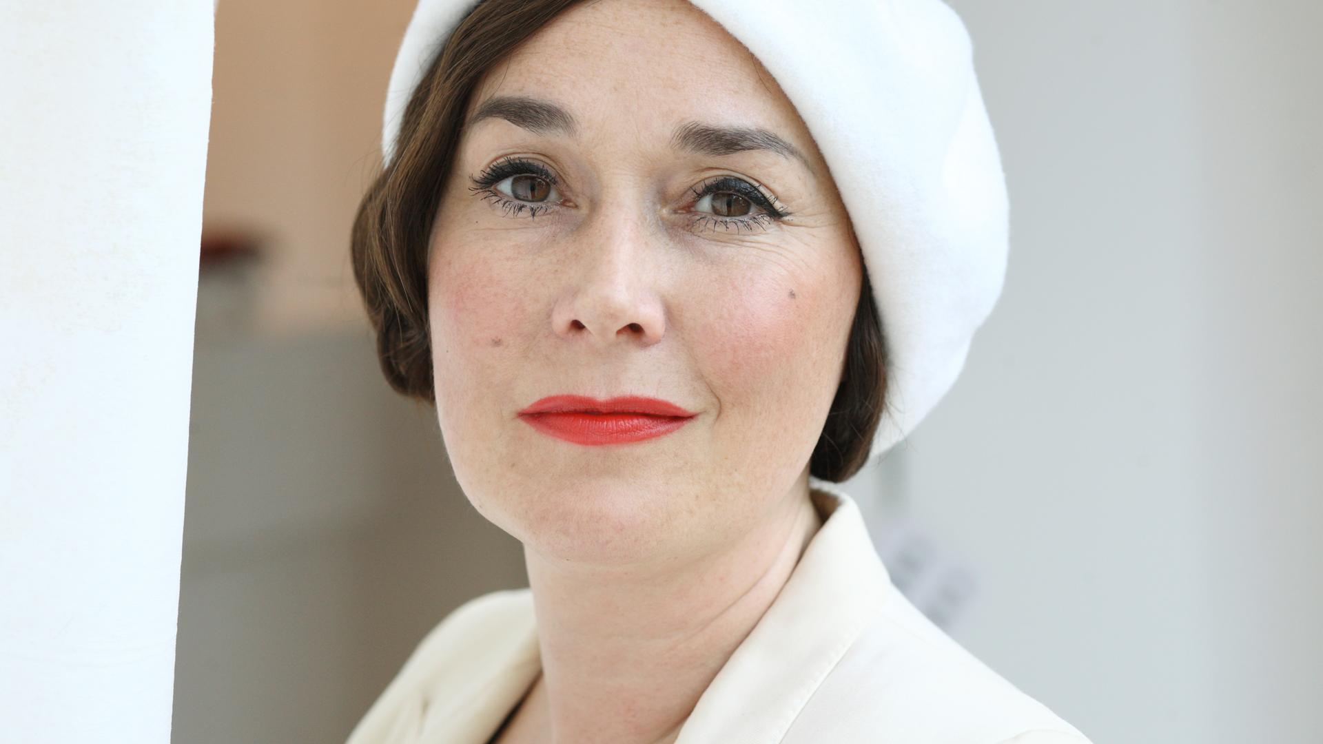 Porträtfoto einer weißen Frau mit kurzen, braunen Haaren, braunen Augen und rot geschminkten Lippen. Sie trägt eine elegante weiße Mütze, die leicht schräg auf ihrem Kopf sitzt. Es handelt sich um Fiona Bennett.