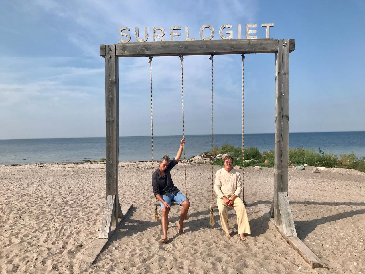 Zwei Männer sitzen am Strand auf den Sitzen einer riesigen Holzschaukel nebeneinander. Zwei Männer sitzen am Strand auf den Sitzen einer riesigen Holzschaukel nebeneinander.