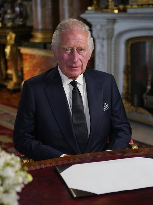 Charles III. sitzt im Londoner Buckingham Palace an einem Schreibtisch, dahinter sieht an einen Kamin