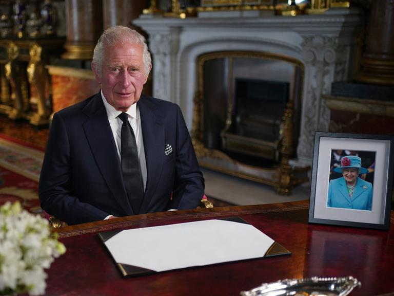 Charles III. sitzt im Londoner Buckingham Palace an einem Schreibtisch, dahinter sieht an einen Kamin