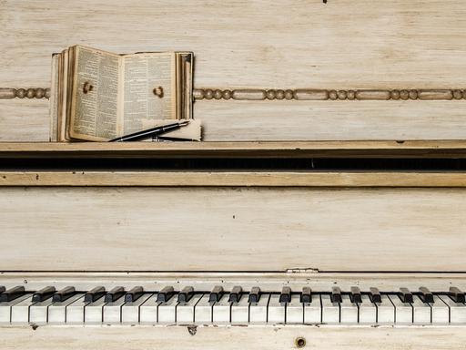 Musik hat eine besondere Anziehungskraft auf Schriftsteller und Autorinnen. Zu sehen ist ein Klavier mit heruntergespielten Tasten, auf der Ablage liegt eine Bibel oder ein ähnliches Buch mit klein geschriebenem Text. 