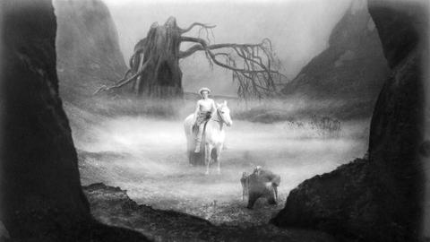 Schwarz-Weiß-Aufnahme: Der oberkörperfreie Siegfried reitet auf einem weißen Pferd durch eine nebelverhangene Wiese. Vor ihm läuft der zwergige Alberich, im Hintergrund steht ein verdorrter Baum.