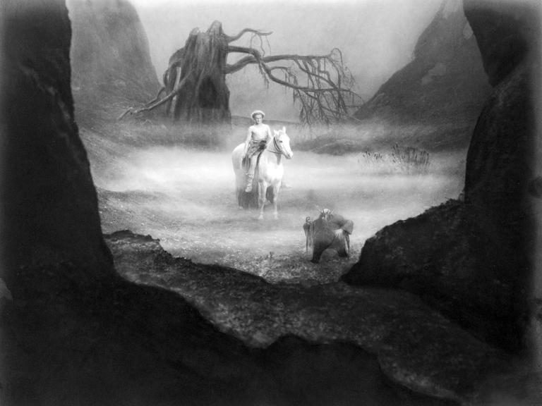 Schwarz-Weiß-Aufnahme: Der oberkörperfreie Siegfried reitet auf einem weißen Pferd durch eine nebelverhangene Wiese. Vor ihm läuft der zwergige Alberich, im Hintergrund steht ein verdorrter Baum.