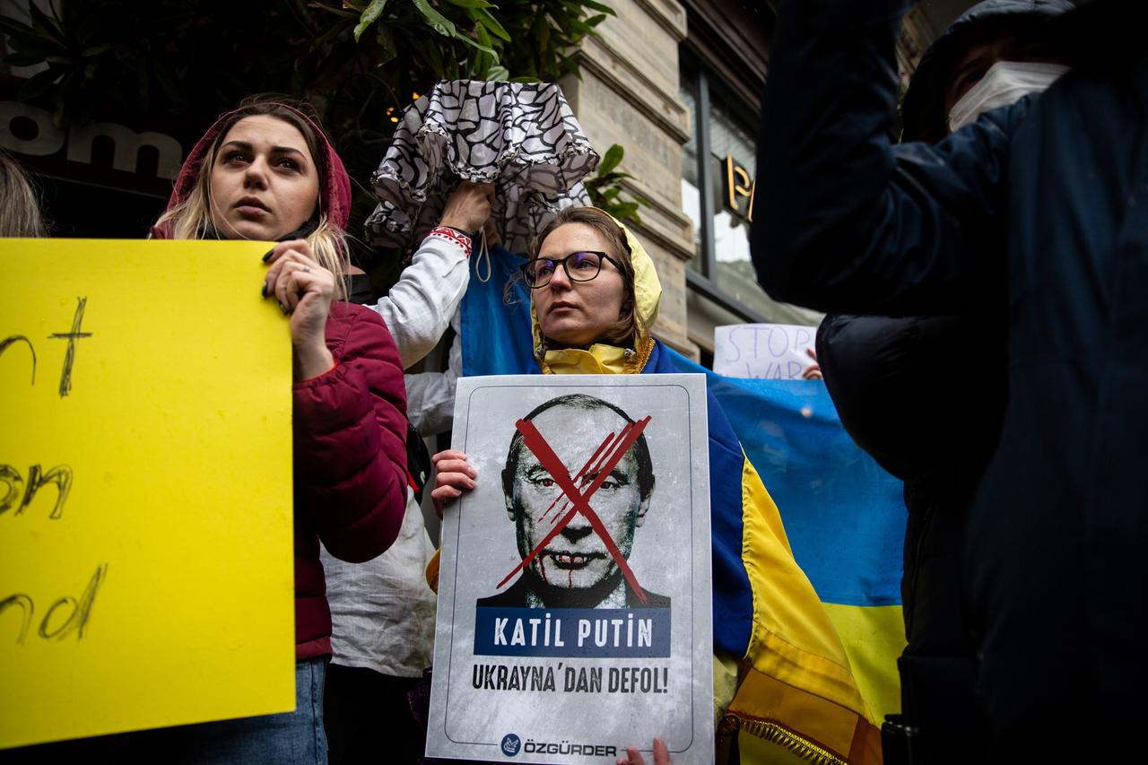 Eine Demonstrantin in gelbblauer Regenjacke trägt ein Porträt von Putin vor sich her, das mit roter Farbe durchgestrichen ist.