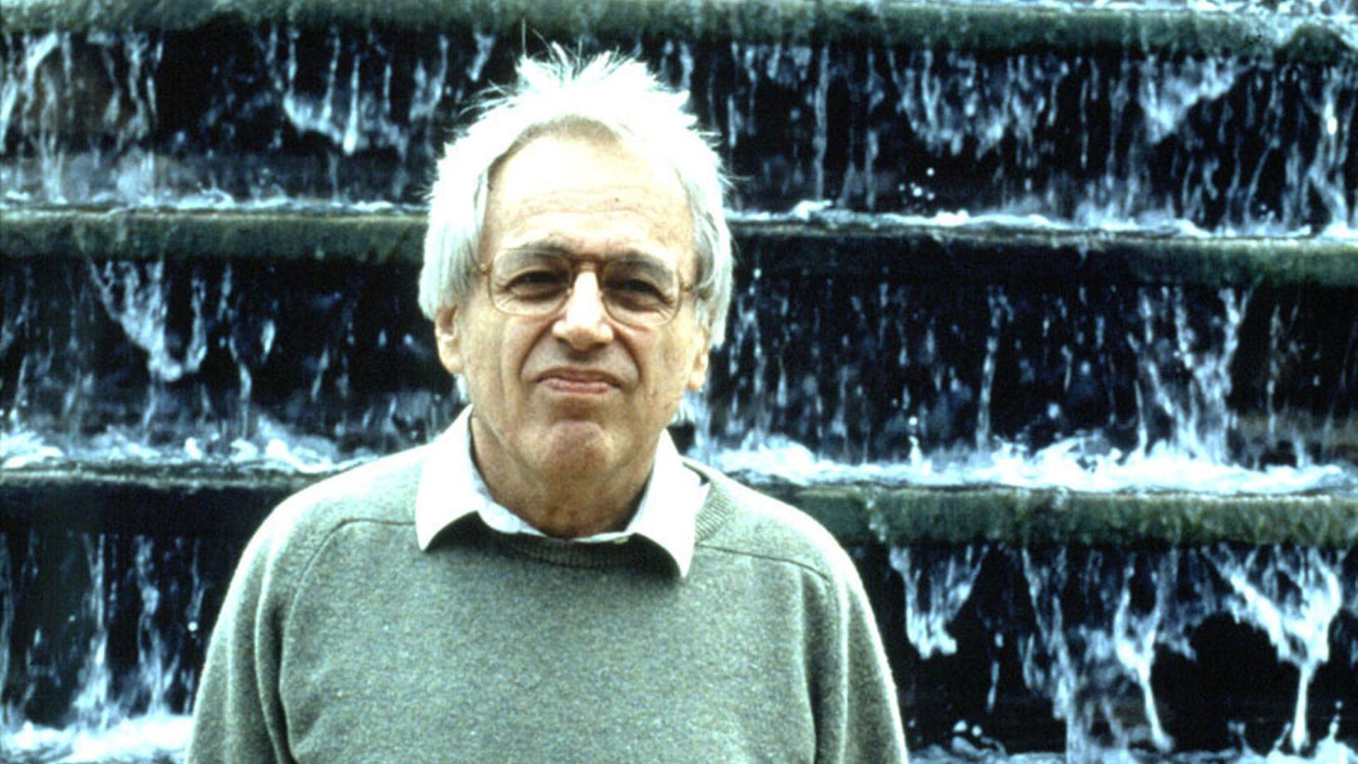 György Ligeti steht mit einem grünlichen Pullover und weißem Haar vor einer Wasserkunst, die wie ein großer Wasserfall aussieht. György Ligeti steht mit einem grünlichen Pullover und weißem Haar vor einer Wasserkunst, die wie ein großer Wasserfall aussieht.