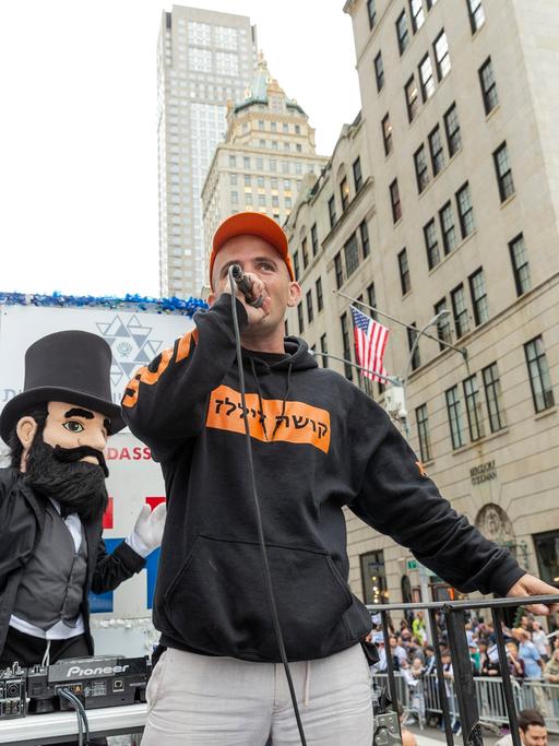 Mann mit Mikrofon auf einem Festwagen, dahinter israelische Flaggen und die Hochhäuser von New York: Der US-Rapper Kosha Dillz bei der Celebrate Israel Parade in New York 2018.