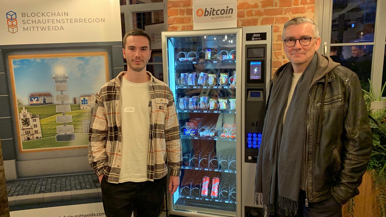 Tim Käbisch, Informatikstudent an der Hochschule Mitttweida, und Andreas Ittner, Informatikprofessor an der Hochschule Mittweida, stehen neben einem Snackautomaten, an dem man mit Bitcoins bezahlen kann.