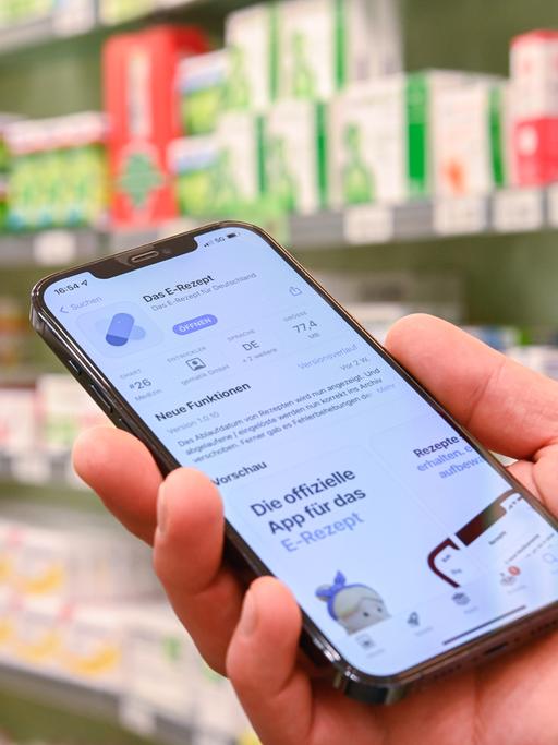 Jemand hält in einer Apotheke ein Smartphone in der Hand, auf dessen Display eine App zur Verarbeitung von E-Rezepten geöffnet ist.