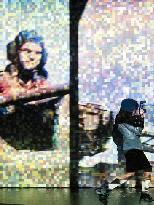 Foto von der Inszenierung: Eine Frau fotografiert im Vordergrund. Im Hintergrund ist ein verpixeltes Bild eines Panzerfahres in Aktion zu sehen.