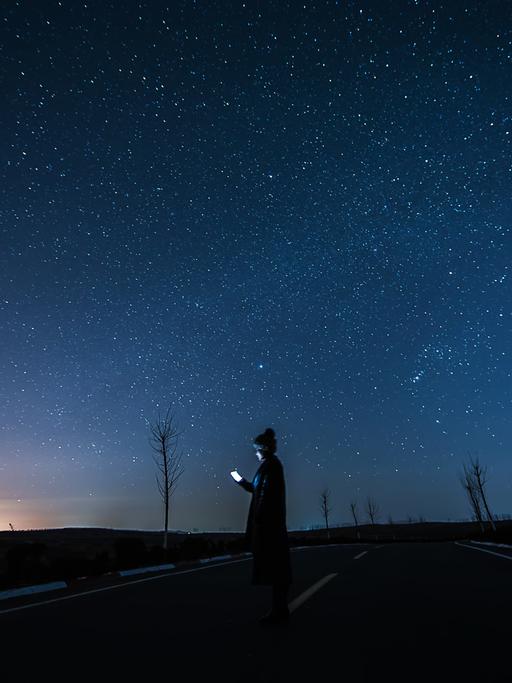 Eine junge Frau steht auf einer einsamen Strasse, ihr Gesicht wird vom Smartphone beleuchtet, darüber ein spektakulärer Sternenhimmel.