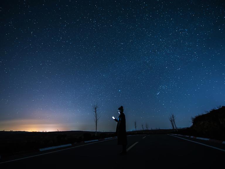 Eine junge Frau steht auf einer einsamen Strasse, ihr Gesicht wird vom Smartphone beleuchtet, darüber ein spektakulärer Sternenhimmel.
