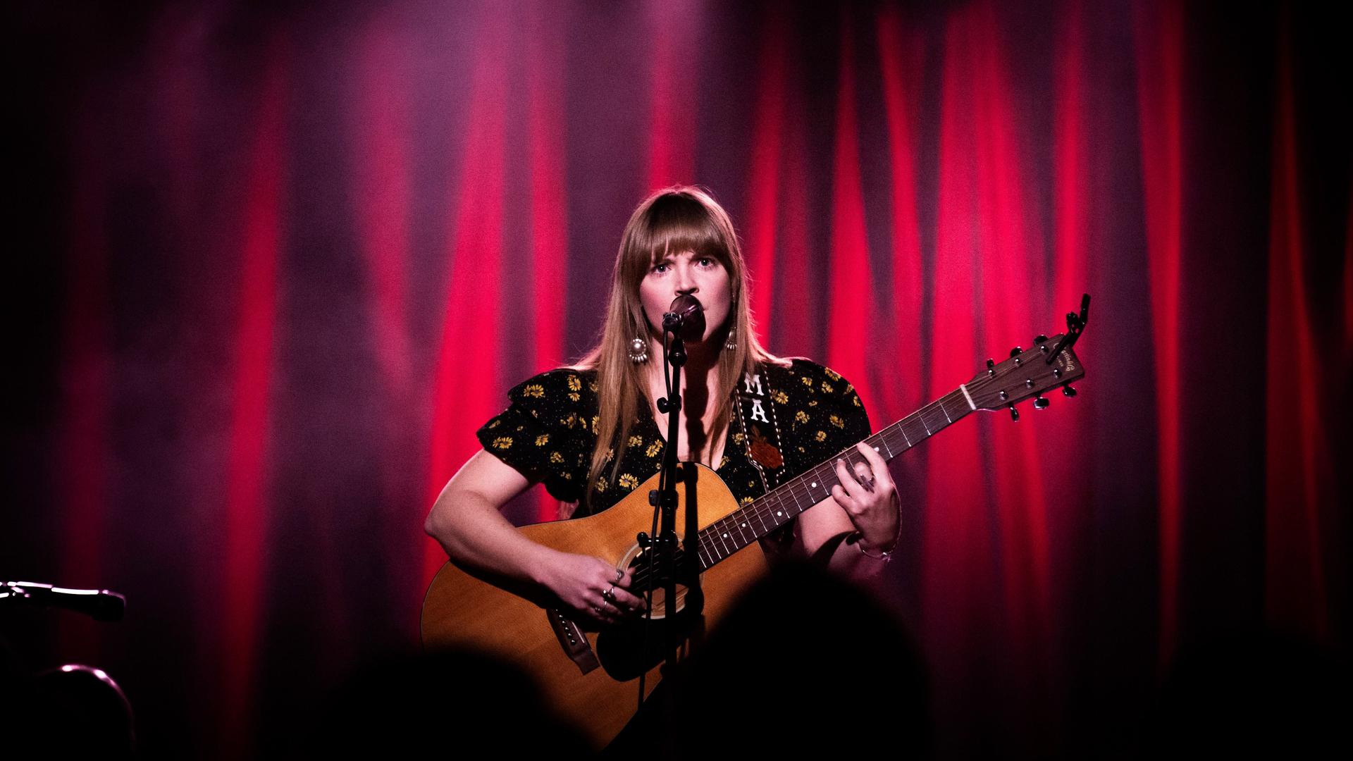 Auf dem Bild ist eine Frau zu sehen, die Gitarre spielt und in ein Mikrofon singt. Auf dem Bild ist eine Frau zu sehen, die Gitarre spielt und in ein Mikrofon singt.