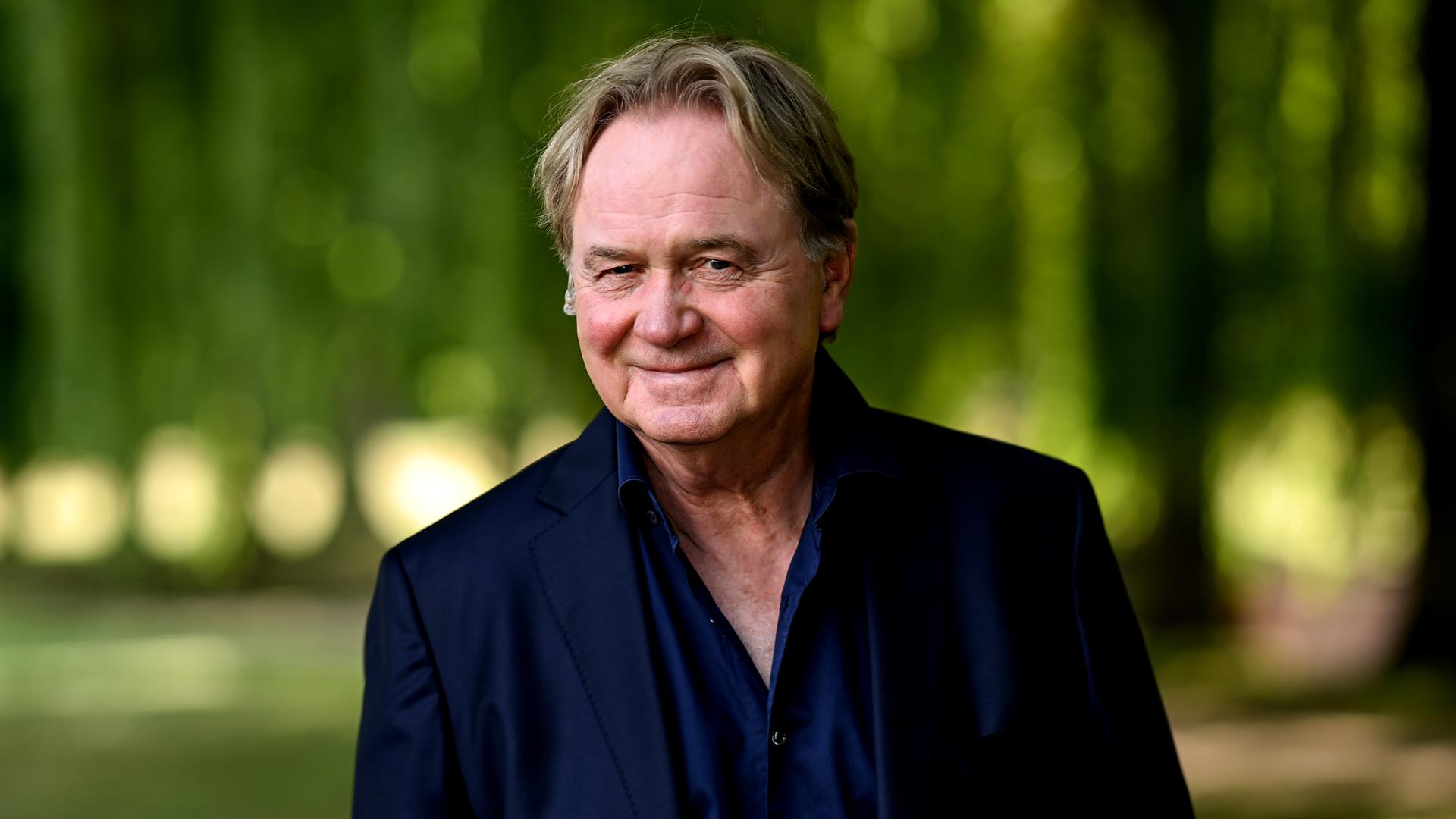 Der Sänger Klaus Hoffmann steht im Grünen und blickt in die Kamera. Er trägt ein blaues Jackett. Der Sänger Klaus Hoffmann steht im Grünen und blickt in die Kamera. Er trägt ein blaues Jackett.