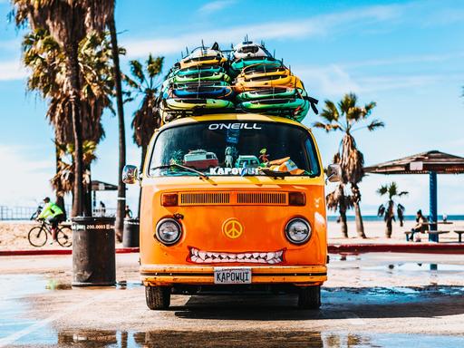 Ein orangfarbener VW Bus mit Surfboards auf dem Dach und einem Peace-Zeichen anstelle des VW-Emblems.