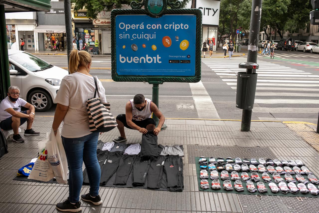 Ein Straßenhändler hockt auf der Straße und verkauft Hosen und Socken. Hinter ihm steht ein Schild mit Bitcoin-Werbung. Ein Straßenhändler hockt auf der Straße und verkauft Hosen und Socken. Hinter ihm steht ein Schild mit Bitcoin-Werbung.