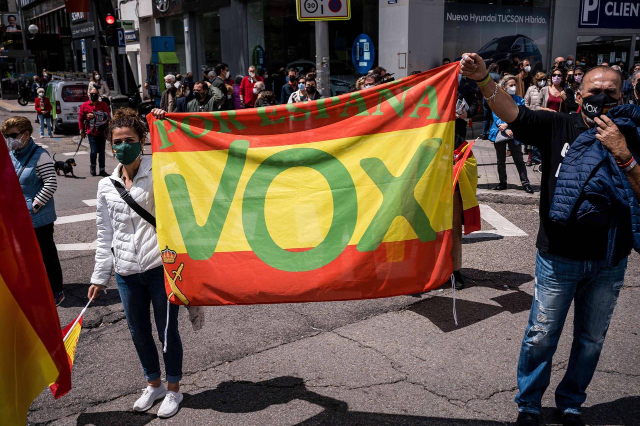Mitten auf der Straße halten zwei Demonstranten zwischen sich ein großes buntes Vox-Plakat in die Höhe.