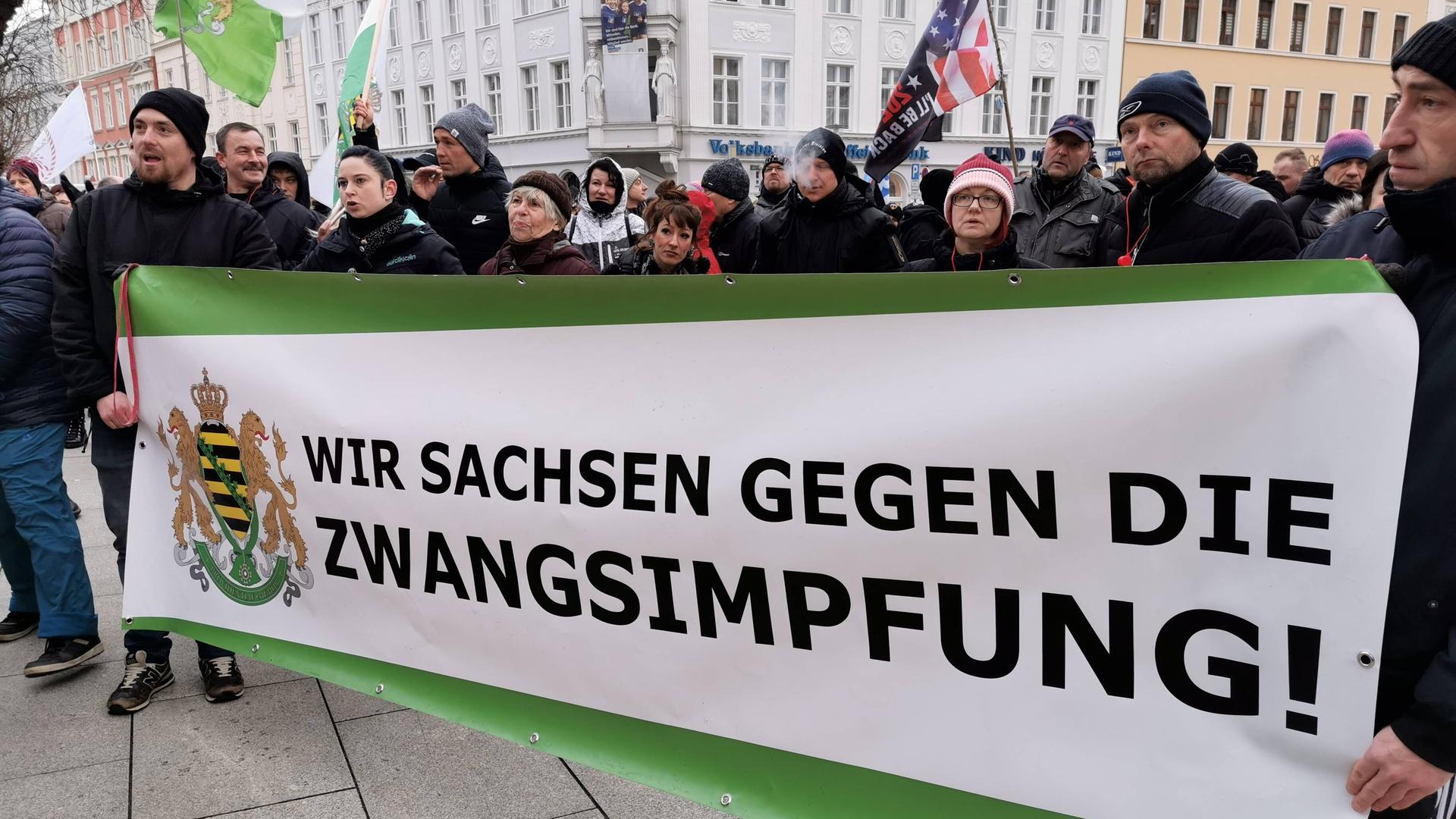 Teilnehmer einer Demonstration halten ein Banner mit der Aufschrift "Wir Sachsen gegen die Zwangsimpfung!", daneben ist das Wappen des Bundeslandes Sachsen abgebildet. 