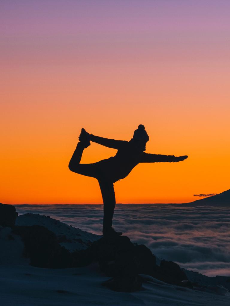 Eine junge Frau macht in Neuseeland Yogaübungen bei Sonnenuntergang über den Wolken, im Hintergrund ist der Mount Taranaki zu sehen.

