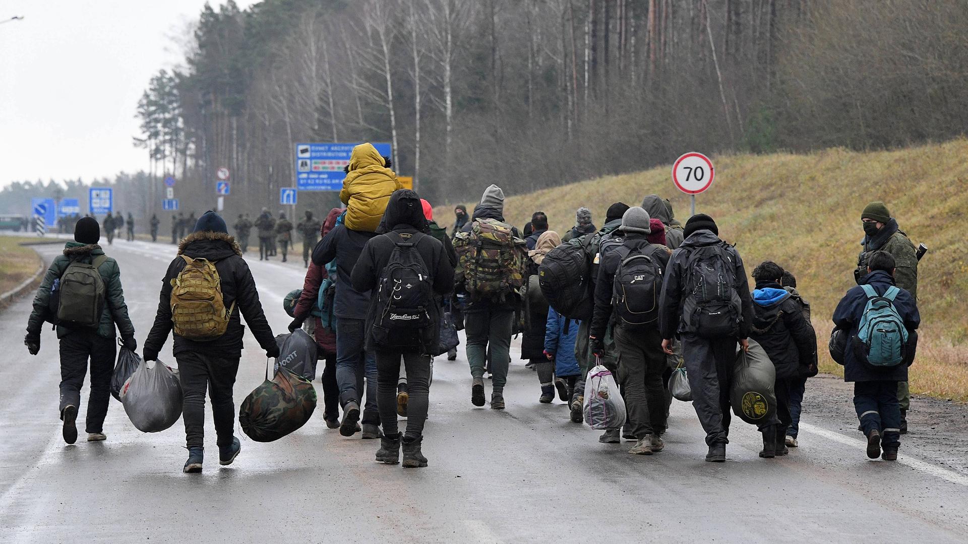 Das Bild zeigt die Gruppe von schwerbepackten Migranten auf einer Straße von hinten. Sie tragen dunkle Kleidung, die Strasse ist feucht. 