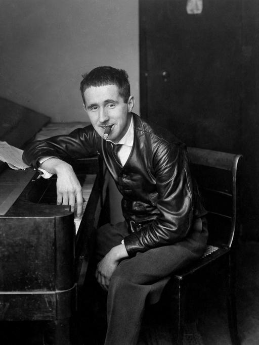 Historische Aufnahme um 1927. Bertolt Brecht sitzt mit Zigarre im Mund an einem Klavier und schaut selbstbewußt Richtung Betrachter.