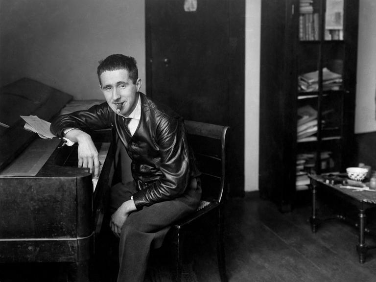 Historische Aufnahme um 1927. Bertolt Brecht sitzt mit Zigarre im Mund an einem Klavier und schaut selbstbewußt Richtung Betrachter.