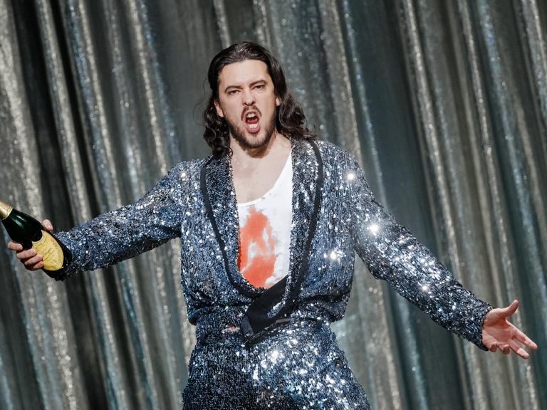 Ein Mann mit langen braunen Locken posiert in Glitzeroutfit und mit Champagnerflasche in der Hand auf einer Bühne. Auf seinem weißen Unterhemd ist Kunstblut zu sehen.