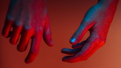 Zwei Hände, die blau, lila und rot beleuchtet sind, hängen von oben herab.