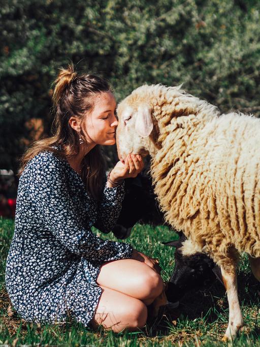 Geraldine Schüle sitzt in einem gepunkteten Kleid auf einer Wiese und küsst ein Schaf.