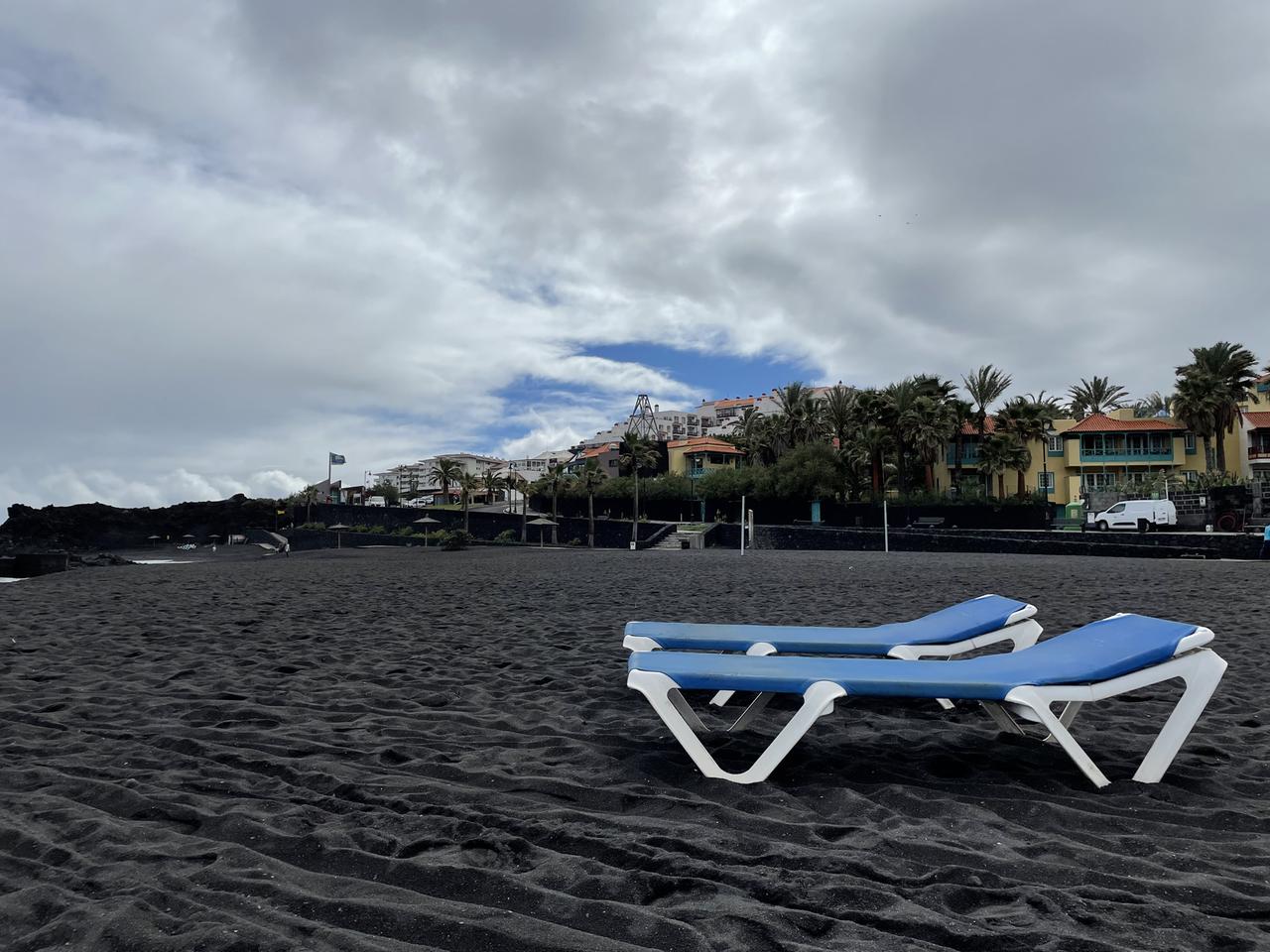 Zwei blaue leere Liegestühle an einem leeren Strand mit schwarzem Sand. Zwei blaue leere Liegestühle an einem leeren Strand mit schwarzem Sand.
