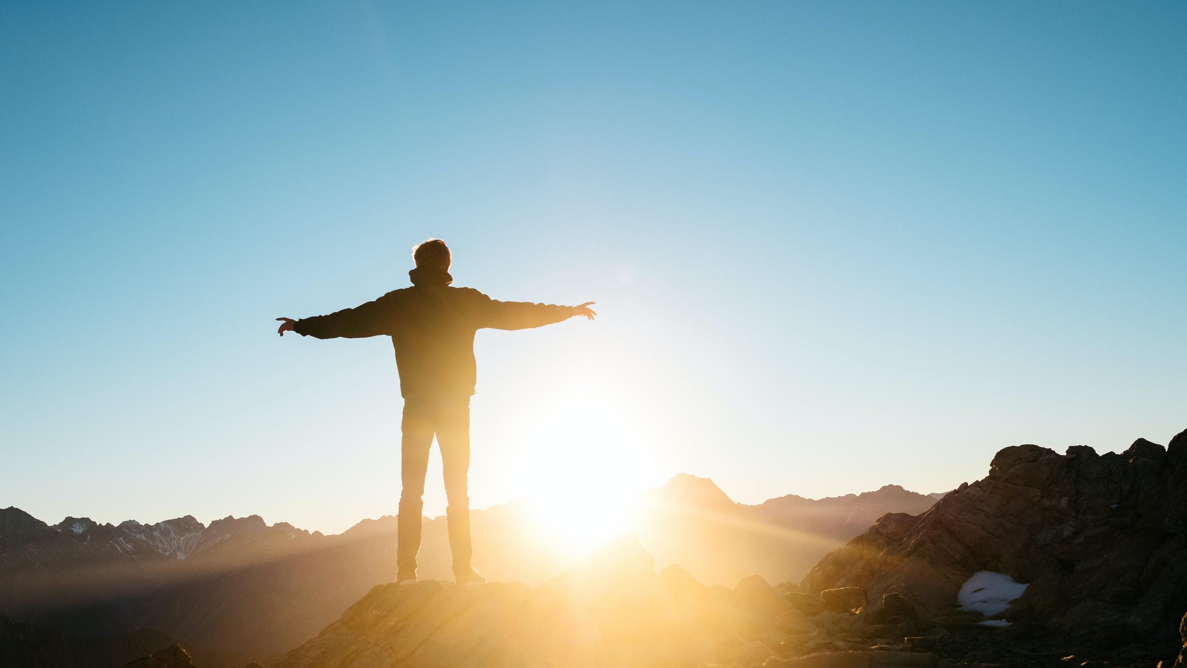 Ein Mann mit ausgebreiteten Armen steht auf einem Berggipfel und schaut der Sonne entgegen.