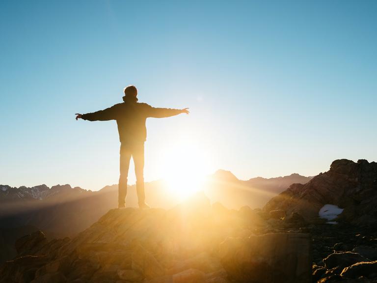 Ein Mann mit ausgebreiteten Armen steht auf einem Berggipfel und schaut der Sonne entgegen.