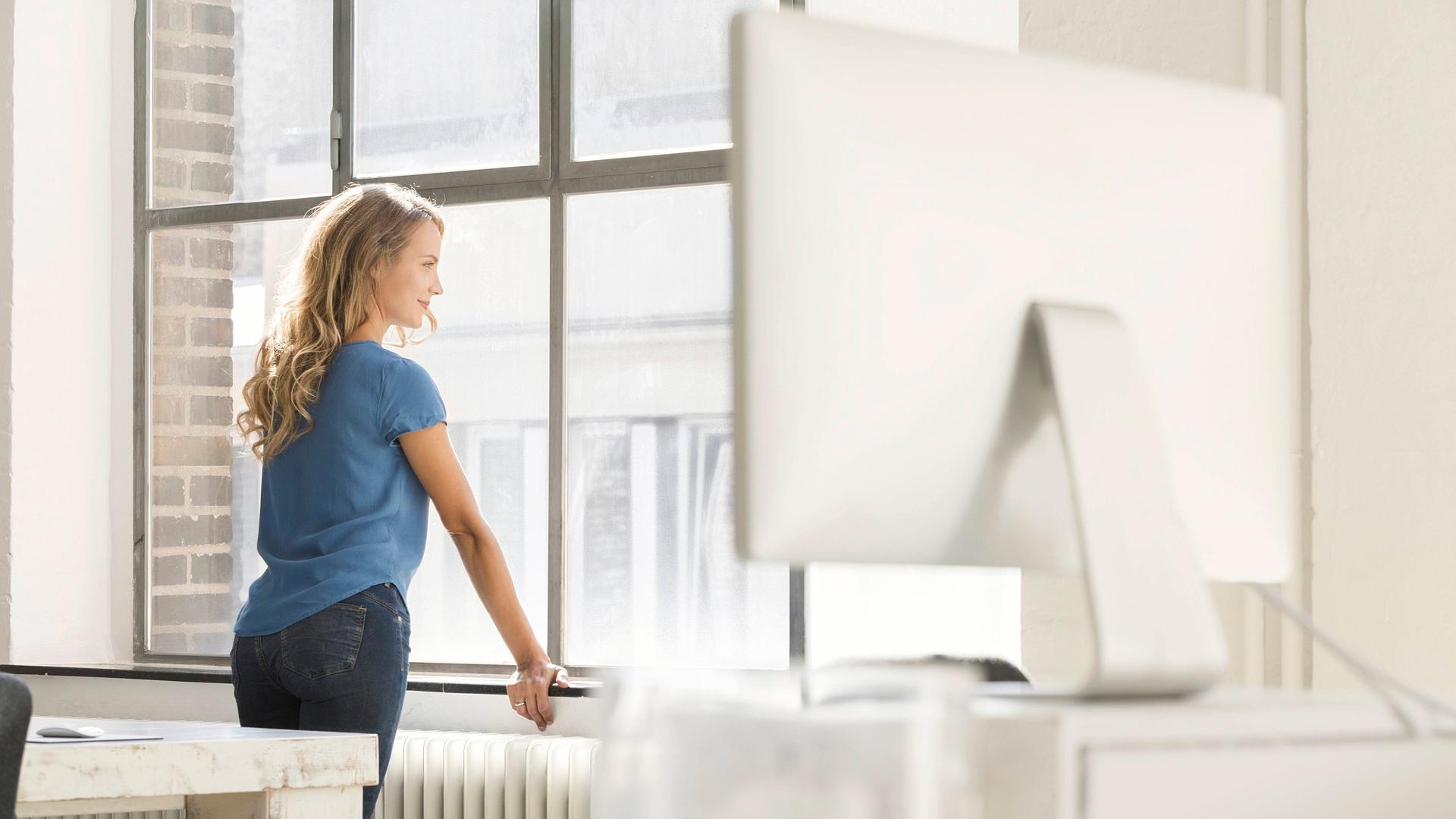 Ein junge blonde Frau mit langen Haaren und hellblauem T-Shirt steht in einem fabriketagen-ähnlichem Büro und schaut aus dem Fenster. Ein junge blonde Frau mit langen Haaren und hellblauem T-Shirt steht in einem fabriketagen-ähnlichem Büro und schaut aus dem Fenster.