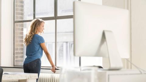 Ein junge blonde Frau mit langen Haaren und hellblauem T-Shirt steht in einem fabriketagen-ähnlichem Büro und schaut aus dem Fenster.
