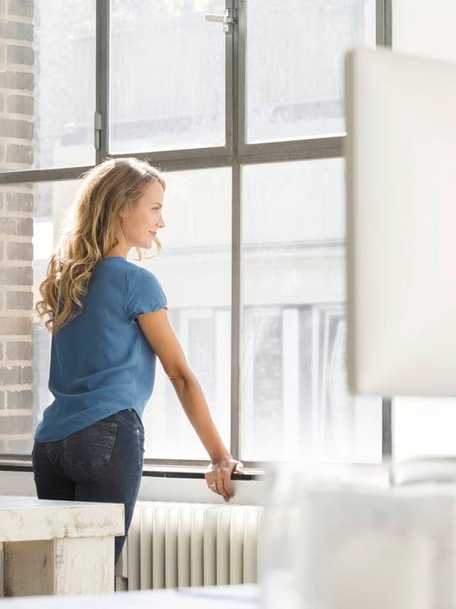 Ein junge blonde Frau mit langen Haaren und hellblauem T-Shirt steht in einem fabriketagen-ähnlichem Büro und schaut aus dem Fenster.