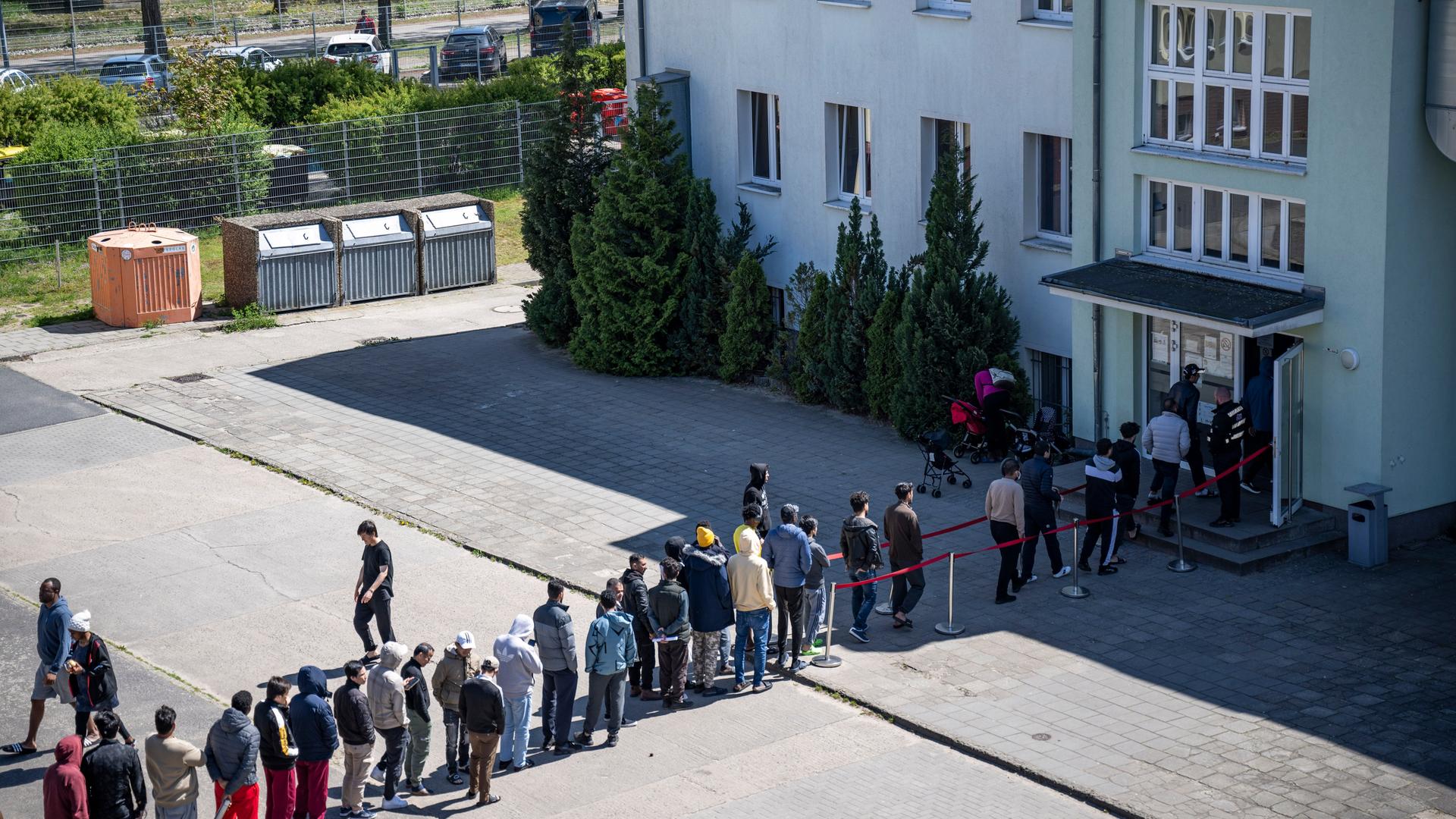 Menschen stehen in einer langen Schlange auf einem Hof vor einem Gebäude und warten auf eine Aufnahme in eine Einrichtung für Geflüchtete. Menschen stehen in einer langen Schlange auf einem Hof vor einem Gebäude und warten auf eine Aufnahme in eine Einrichtung für Geflüchtete.