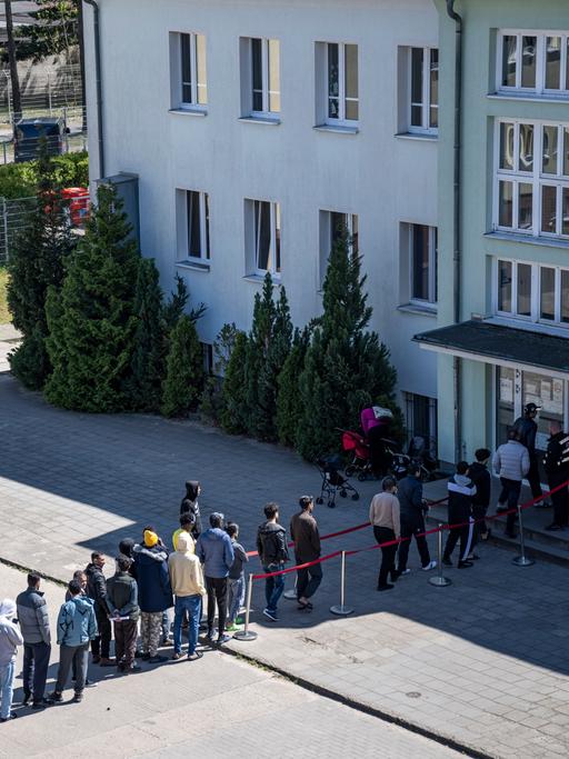 Menschen stehen in einer langen Schlange auf einem Hof vor einem Gebäude und warten auf eine Aufnahme in eine Einrichtung für Geflüchtete.