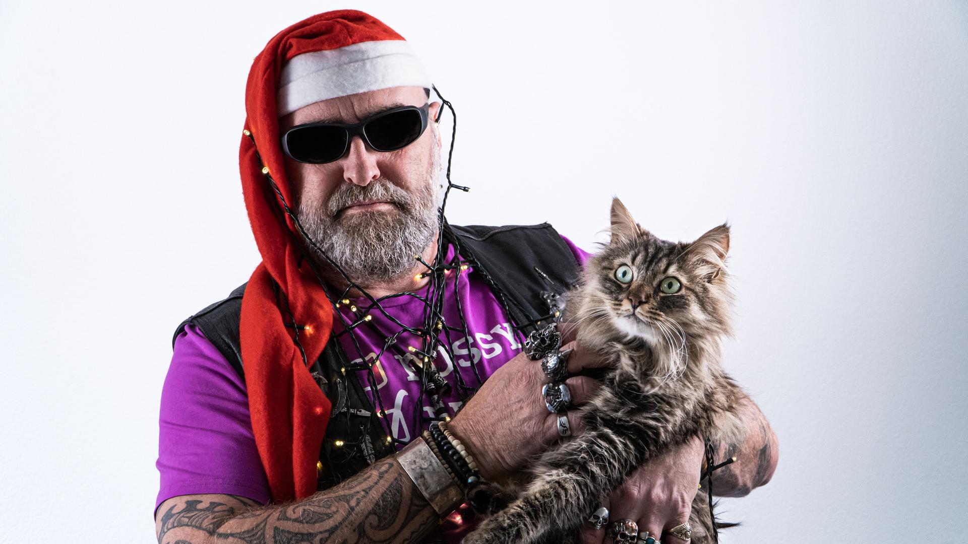 Ein Mann voller Tattoos und Vollbart trägt Weihnachtsmannmütze und Brille, eine Weihnachtsbaumlichterkette um Kopf und Hals gewickelt sowie eine Katze auf dem Arm, die er grault. Ein Mann voller Tattoos und Vollbart trägt Weihnachtsmannmütze und Brille, eine Weihnachtsbaumlichterkette um Kopf und Hals gewickelt sowie eine Katze auf dem Arm, die er grault.