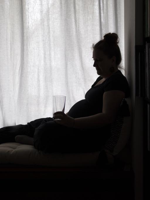 Eine Frau sitzt erschöpft an einem Fenster, in der Hand ein Wasserglas.