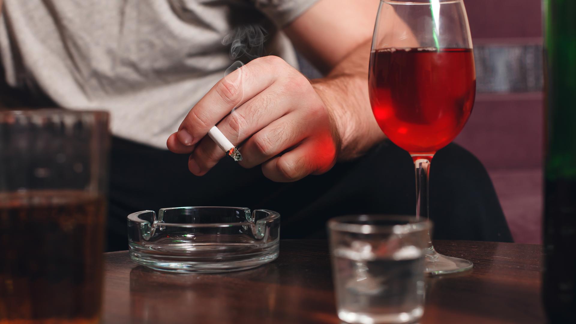 Hand eines Mannes hält eine Zigarette, daneben steht ein Rotwein-Glas. (Symbolfoto) Hand eines Mannes hält eine Zigarette, daneben steht ein Rotwein-Glas. (Symbolfoto)