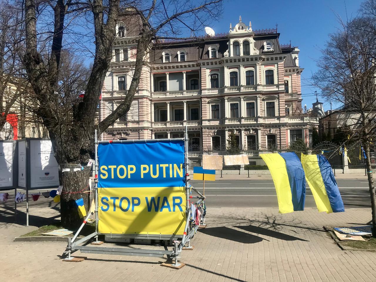 Ein blau-gelbes Plakat mit "Stop Putin, Stop den Krieg" steht an der Straße, gegenüber von einem repräsentativem Gebäude.