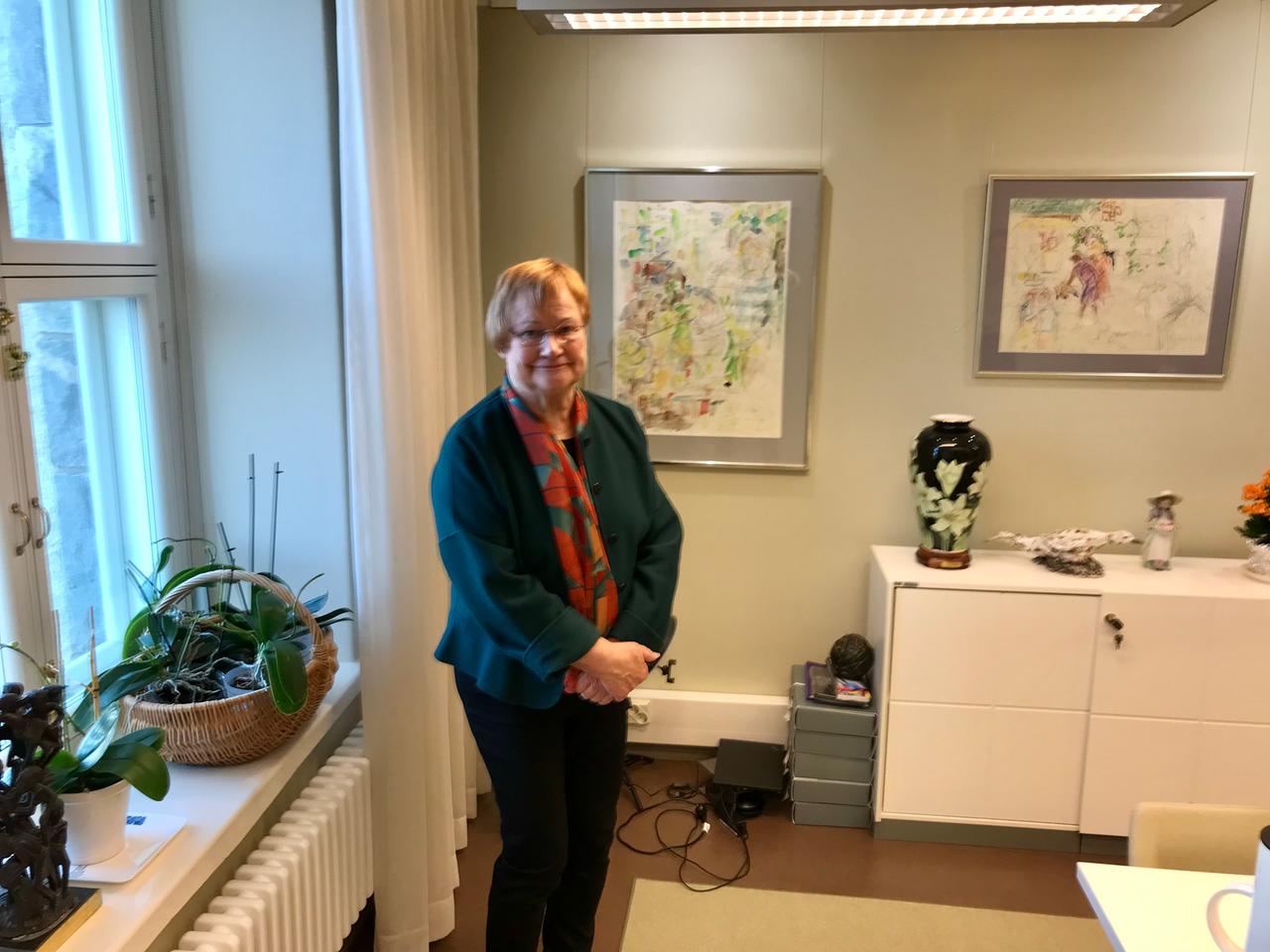 Eine ältere Dame mit roten Haaren, Brille und grünem Blazer steht in einem Wohnzimmer mit Blumen und Vasen. Eine ältere Dame mit roten Haaren, Brille und grünem Blazer steht in einem Wohnzimmer mit Blumen und Vasen.